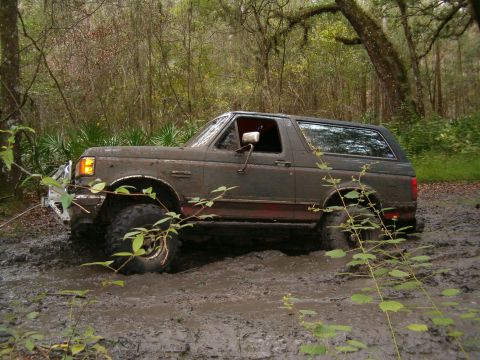 1991 Ford Bronco - Driver-side profile of a lifted Ford Bronco sitting hub-deep in a rutted mud hole on a wooded trail, its body and windows plastered with we…