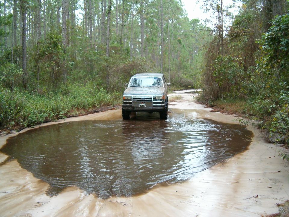 1991 Ford Bronco driving through a large puddle on a dirt road surrounded by trees