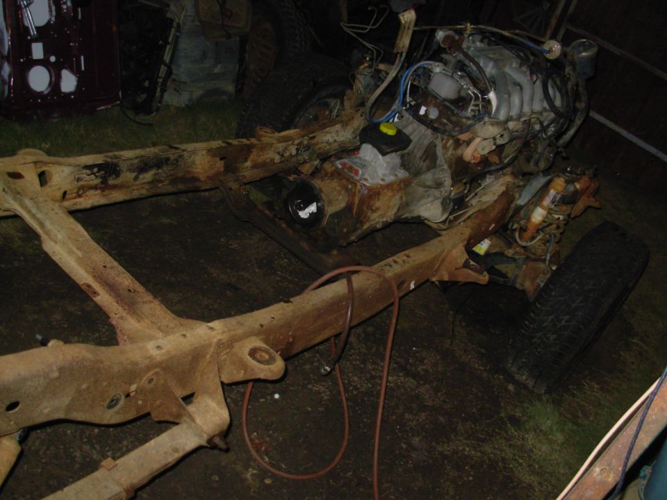 Rusty stripped truck frame with engine and one tire in a dirt-floor shed