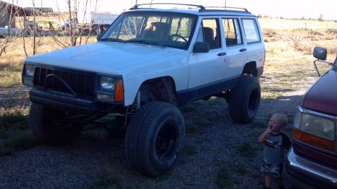 1995 Jeep Cherokee - A white Jeep Cherokee with significant lift and oversized tires sits in a gravel lot next to another vehicle and a small child.