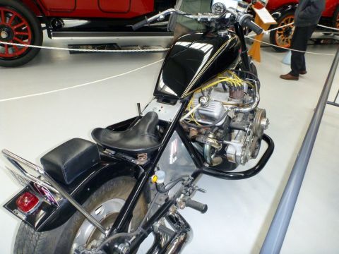 1969 Lincoln Continental - A custom motorcycle with a black fuel tank and exposed engine is displayed indoors, with a vintage red car visible behind it.