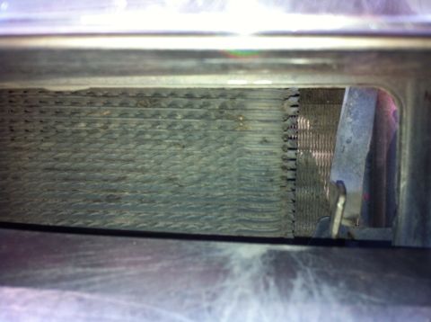1992 Ford F-150 - Tight close-up through a grille opening at a radiator or condenser core, showing bent and dirt-clogged aluminum fins along the upper tank s…
