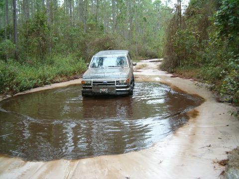 1991 Ford Bronco - A gray Ford Bronco navigates a water-filled dirt road surrounded by trees.