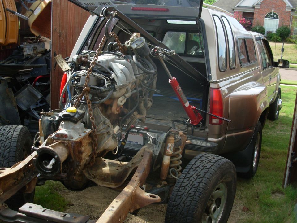 Engine on chain hoist beside tan SUV and bare frame with suspension in a driveway