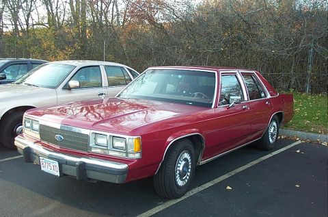 1991 Ford LTD Crown Victoria - A red sedan is parked in a lot next to another vehicle, with trees and a fence in the background.