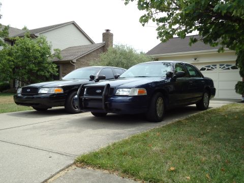 2001 Ford Crown Victoria - Two Ford Crown Victorias, a blue one with a push bar and a black one, are parked in a driveway in front of a garage.