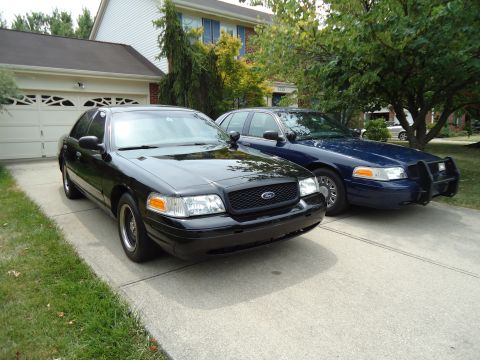 2001 Ford Crown Victoria - Two Ford Crown Victorias, a black civilian model and a blue police interceptor, parked outside a suburban home.
