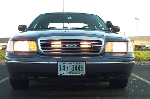 1991 Ford LTD Crown Victoria - Front view of a dark blue sedan with its headlights illuminated, parked in a lot.