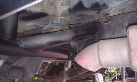 1978 Ford Bronco - Underbody view of a black welded exhaust pipe running beside a frame rail, with a rusty catalytic converter or muffler at right.