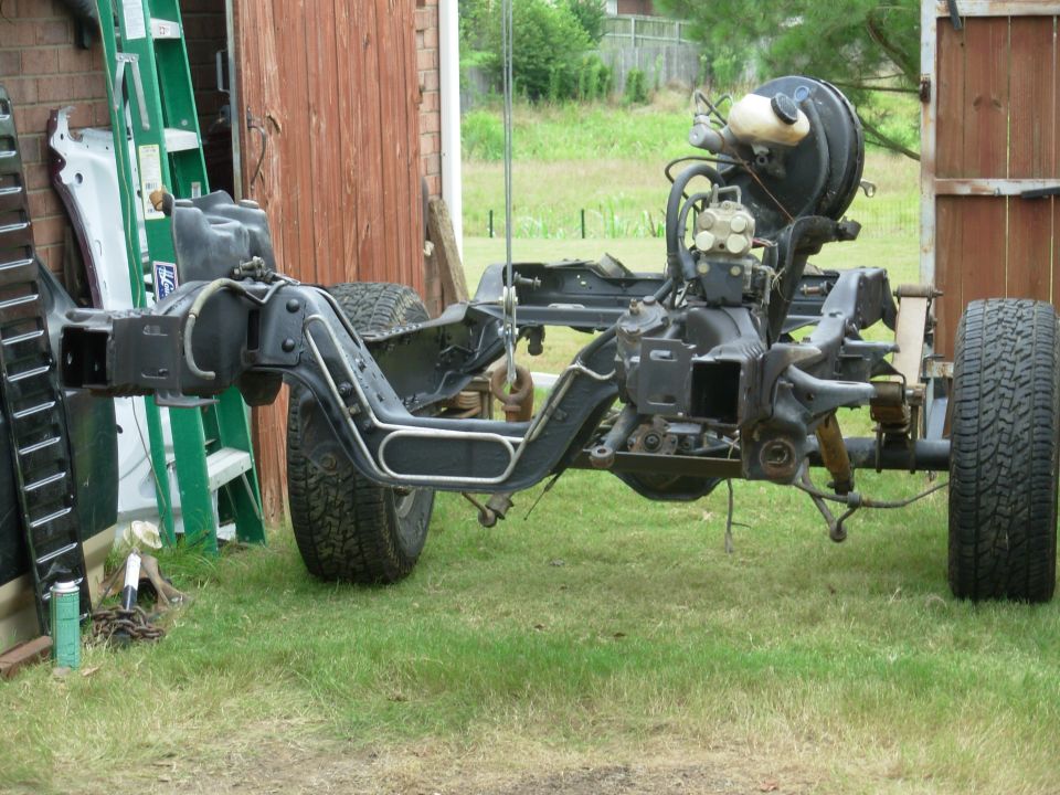 Bare black truck frame with front suspension, wheels and brake booster on grass by a shed