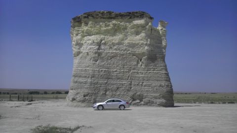 1989 Ford Bronco - A silver sedan is parked in front of a massive rock formation in a wide-open, arid landscape.
