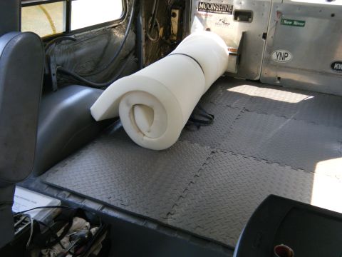 1988 Ford E350 - The interior of a van shows a rolled mattress on gray diamond plate flooring, with various stickers on the rear door.