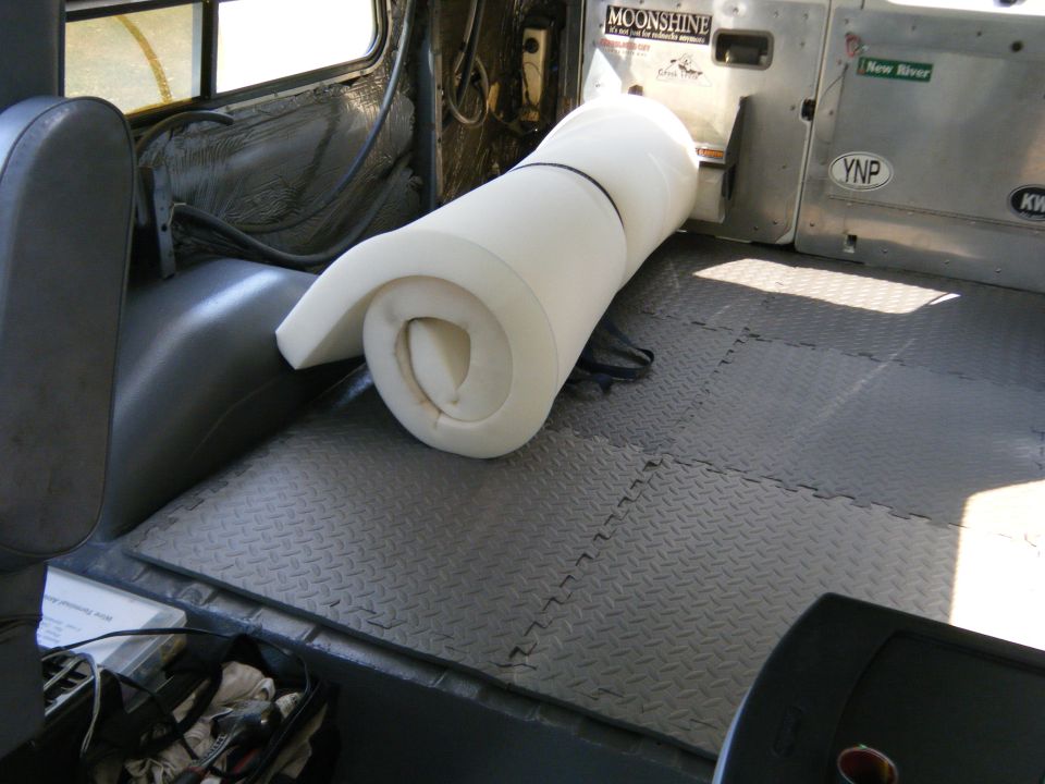 Van interior with gray diamond plate flooring and a rolled white mattress. Stickers on the rear door include 'MOONSHINE' and 'New River'