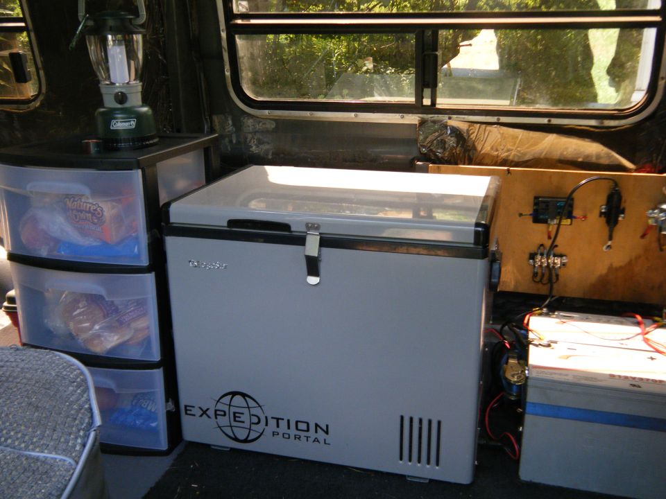 Interior view of a van showing a gray portable refrigerator with "EXPEDITION PORTAL" text, storage drawers, and a battery setup