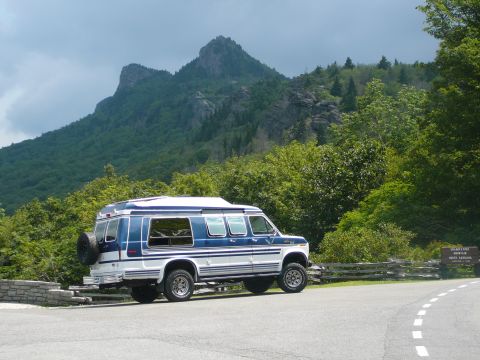 1988 Ford E350 - A blue and white conversion van is parked on the side of a mountain road with a large mountain in the background.