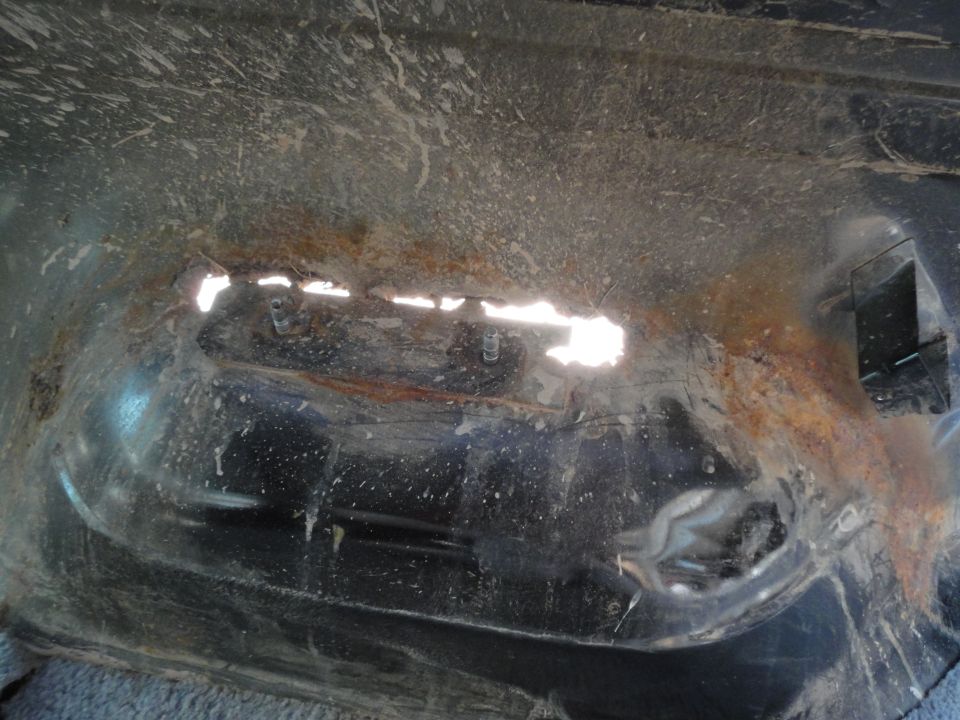 Rusted metal panel with holes letting light through, two studs on a dirty bracket, heavy rust and grime