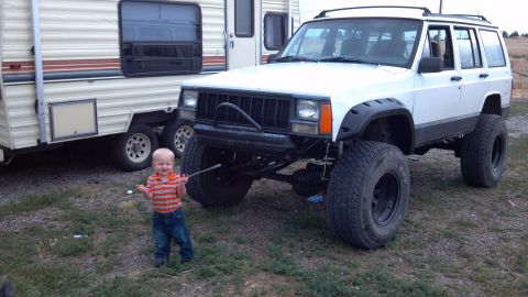 1995 Jeep Cherokee - A white 1995 Jeep Cherokee, modified with large tires, is parked next to a trailer with a young child standing in front of it.