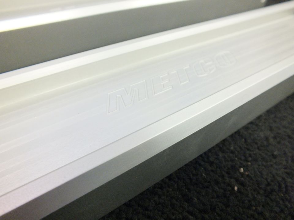 Closeup of billet aluminum suspension control arm with engraved METCO logo on carpeted surface