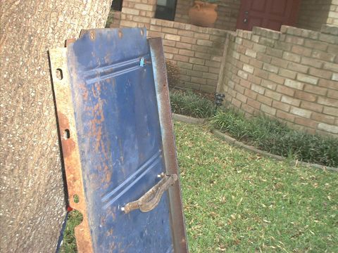 1971 Ford Bronco - A blue tailgate with a chrome handle is shown leaning against a tree, with a brick wall and yard in the background.