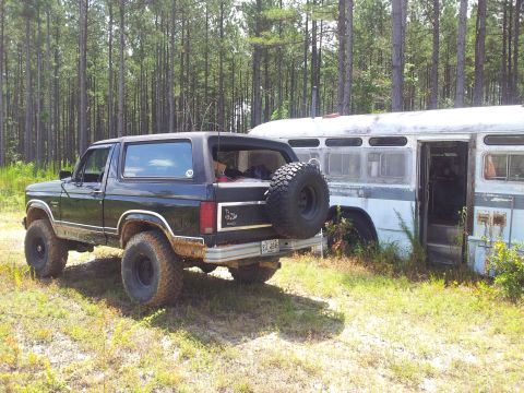 1986 Ford Bronco - A black Ford Bronco is parked next to an old white bus in a grassy, wooded area.