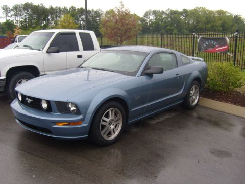 2007 Ford Mustang - A blue 2007 Ford Mustang coupe is parked on a wet asphalt lot next to a white pickup truck.