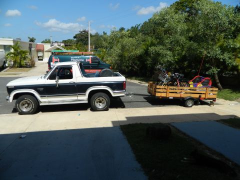 1984 Ford Bronco - A two-tone pickup truck tows a trailer loaded with various items down a driveway.
