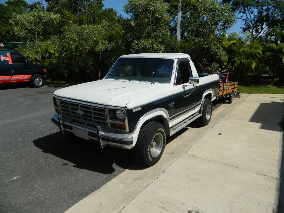 White and dark green Ford F-Series pickup truck with a small wooden trailer attached. Another dark van is parked to the left