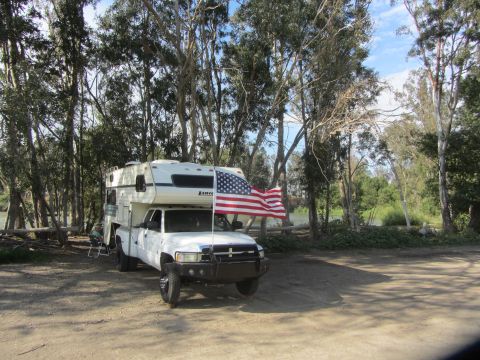 1968 Ford Mustang - A truck camper is parked in a wooded area next to a body of water, with an American flag flying.