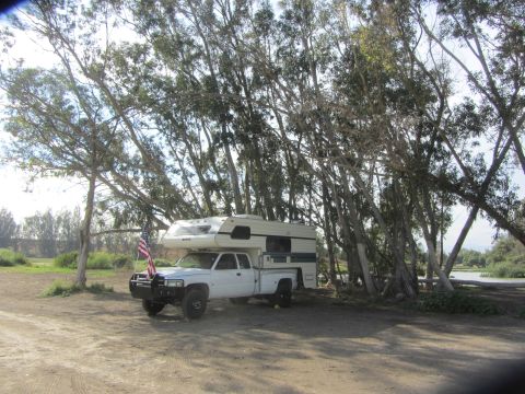 1968 Ford Mustang - A white pickup truck with a camper is parked in a dirt lot near trees and a body of water.