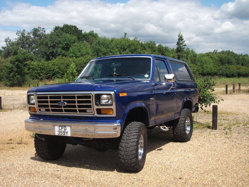 Blue 1982 Ford Bronco parked on gravel with trees and sky in background. License plate CPD 359Y