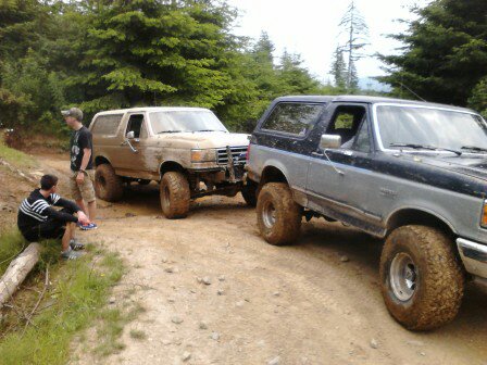 1989 Ford Bronco - Two Ford Broncos are parked on a dirt road, with two people standing near one of them.