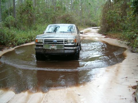 1991 Ford Bronco - A 1987 Ford Bronco navigates a water-filled dirt path surrounded by trees.