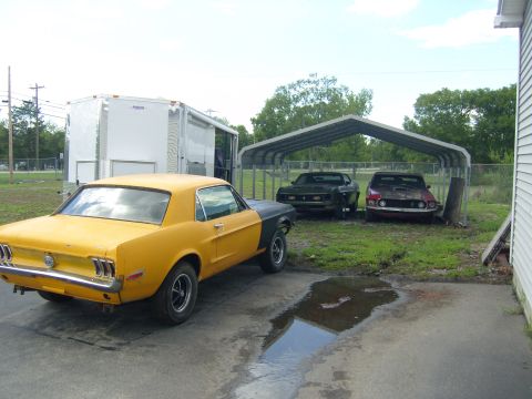 2007 Ford Mustang - Three classic cars are parked outside, two under a metal carport and one yellow and black coupe in the foreground.