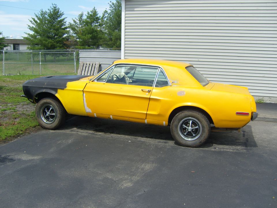 Yellow classic car with a black hood, primer patches on the body, and aftermarket wheels parked on asphalt