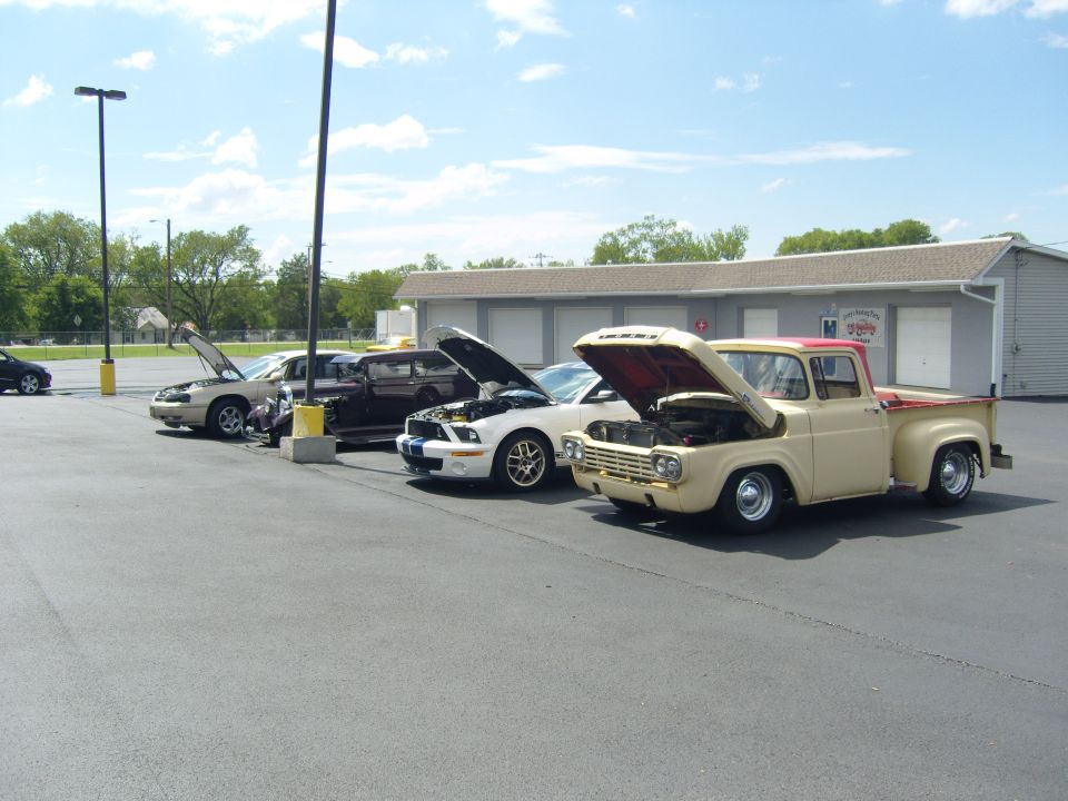Several cars parked in a lot with hoods up, including a tan 1959 Ford pickup and a white Ford Mustang