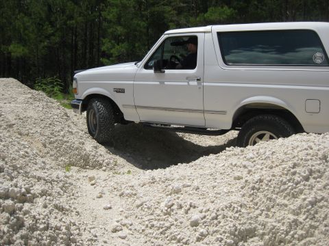 1994 Ford Bronco - A white 1994 Ford Bronco navigates a rough, sandy terrain with trees in the background.