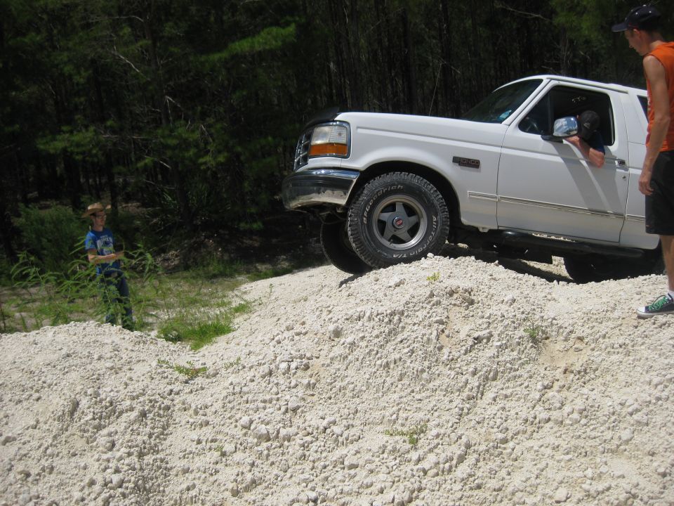 White 1994 Ford Bronco climbing a dirt mound with two people nearby