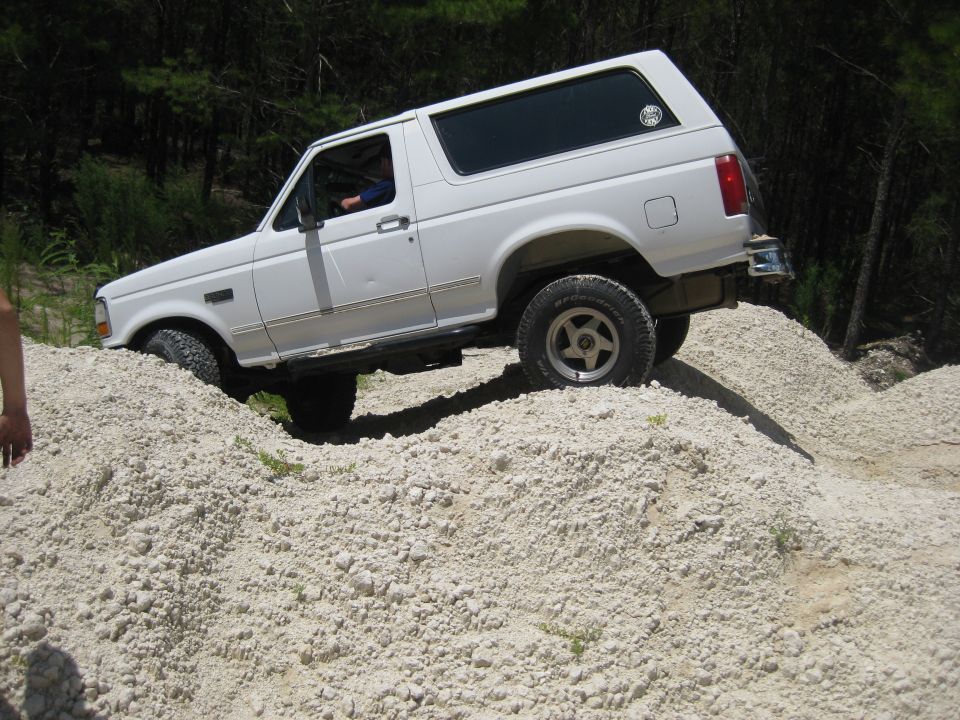 White 1992 Ford Bronco climbing a dirt mound, driver visible inside