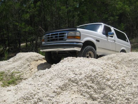 1994 Ford Bronco - A white 1994 Ford Bronco navigates a rough, sandy terrain with trees in the background.