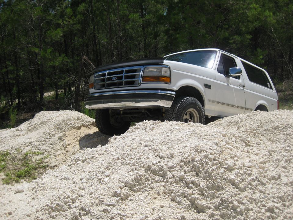White 1994 Ford Bronco driving over a pile of light-colored dirt. Trees are in the background