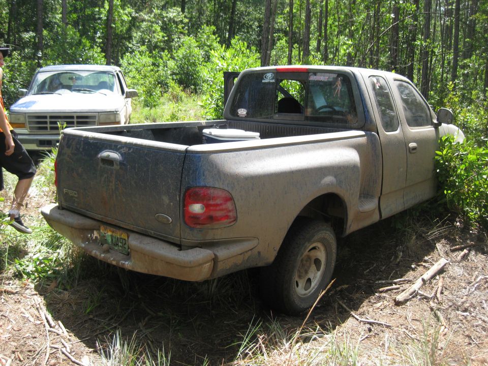Mud-covered 1997 Ford F-150 pickup truck, rear view, parked off-road with another truck behind it