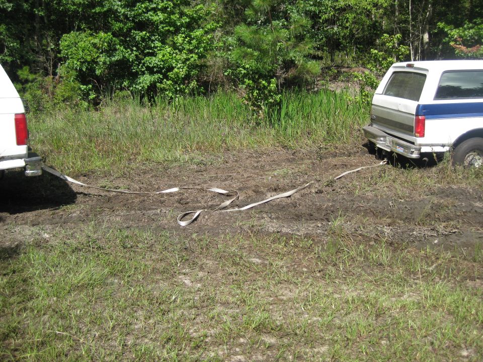 White and blue SUV stuck in mud, being pulled by another vehicle with a tow strap