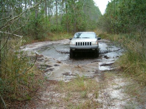1991 Ford Bronco - Silver Jeep Grand Cherokee drives through a muddy, water-filled road in the woods.