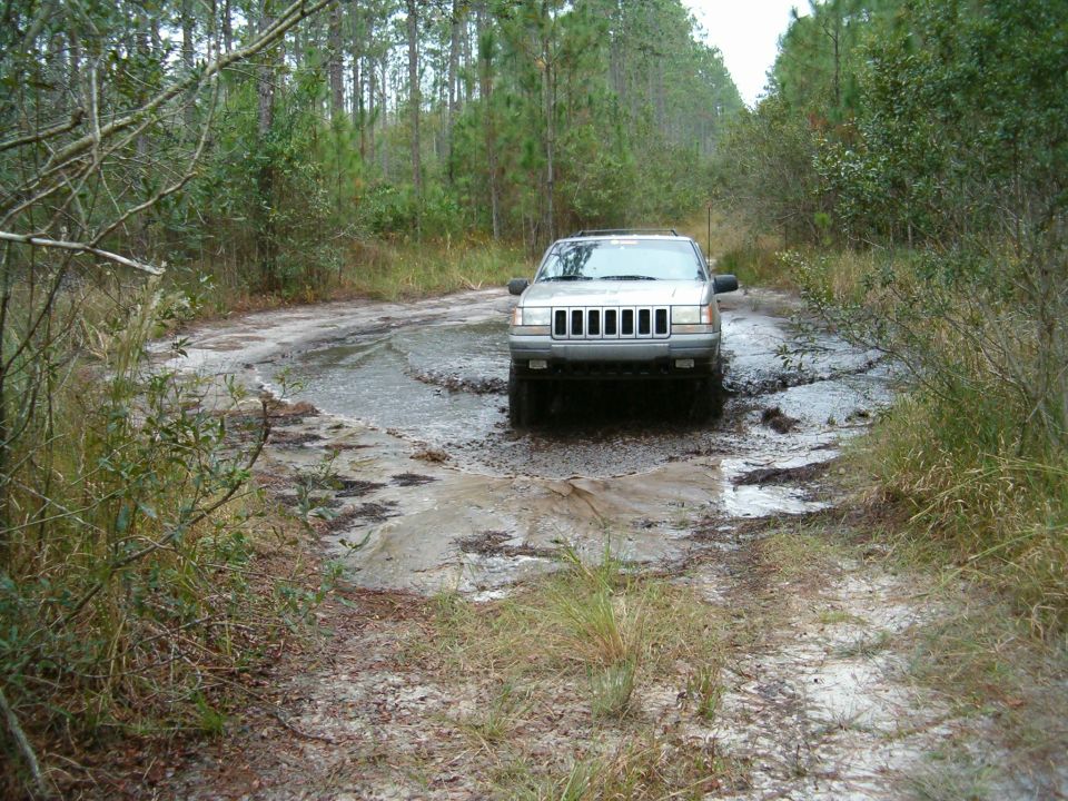 Silver Jeep Grand Cherokee navigating a muddy, pothole-filled dirt road in a wooded area. Water splashes around the tires