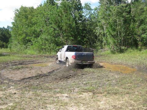 1994 Ford Bronco - A white pickup truck churns through a muddy trail surrounded by trees. The vehicle appears to be from the early 2000s.
