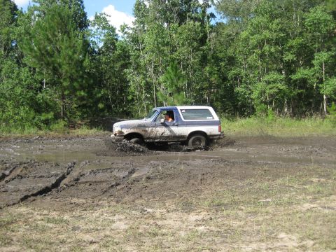 1994 Ford Bronco - A Ford Bronco navigates a challenging muddy path, kicking up dirt and water.