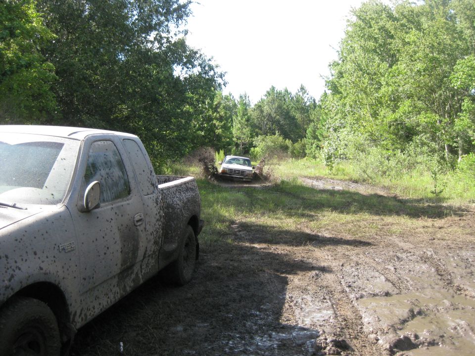Muddy pickup truck in foreground, another truck splashing through mud puddle in background