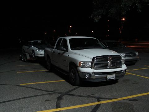 1992 Ford Taurus - A white pickup truck is parked in a lot at night, towing a car on a trailer behind it.