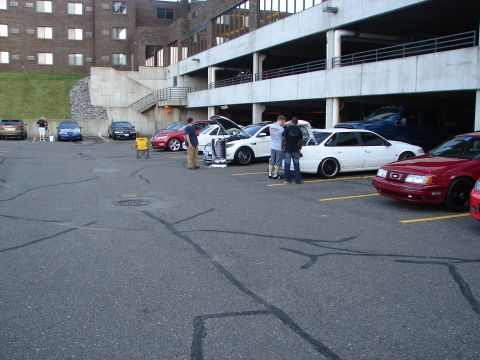 1992 Ford Taurus - A gathering of vehicles and people in a parking lot outside a building.