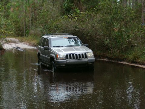 1991 Ford Bronco - A silver Jeep Grand Cherokee navigates through a shallow body of water on a dirt path surrounded by trees.